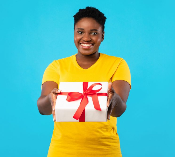 Cheerful Plus Size African Woman Offering Wrapped Gift Box To Camera Posing Standing Over Blue Studio Background. This Present Is For You. Birthday Celebration, Holiday Gifts And Presents Concept